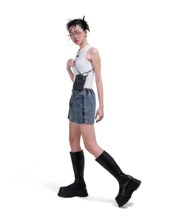 Full-body shot of a female model in urban streetwear featuring a white tank top and boots, showcasing the black SYMA Imperial modular everyday carry wallet worn as a hanging neck pouch