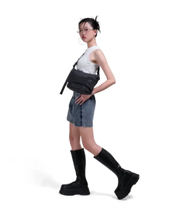 Woman in edgy streetwear outfit posing with the black SYMA Swift worn across her body, demonstrating its compact scale. Utility crossbody sling bag.
