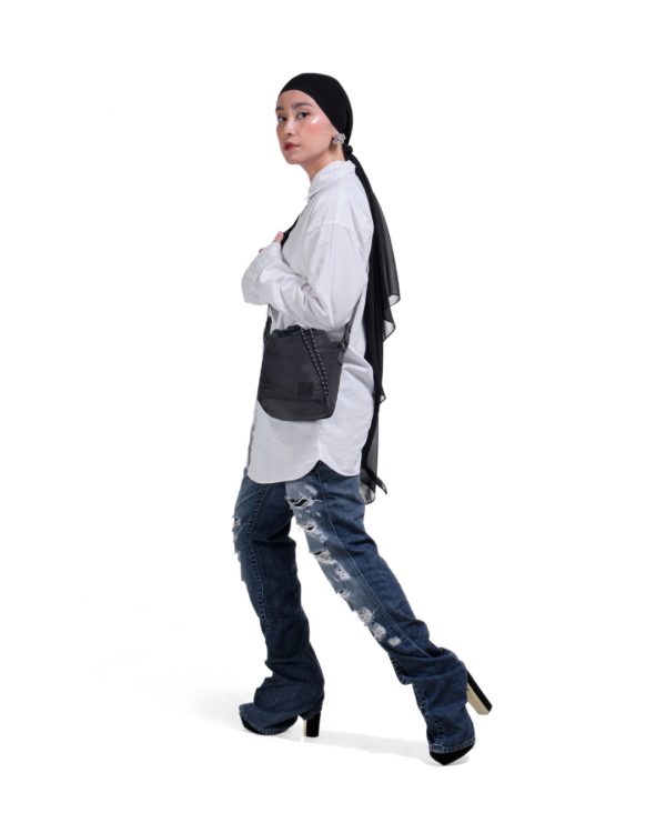 Full-body side view of a female model in modest urban streetwear wearing the black SYMA Sense Sling Bag, demonstrating its compact scale and quick-access closure. Minimalist drawstring sling bag.