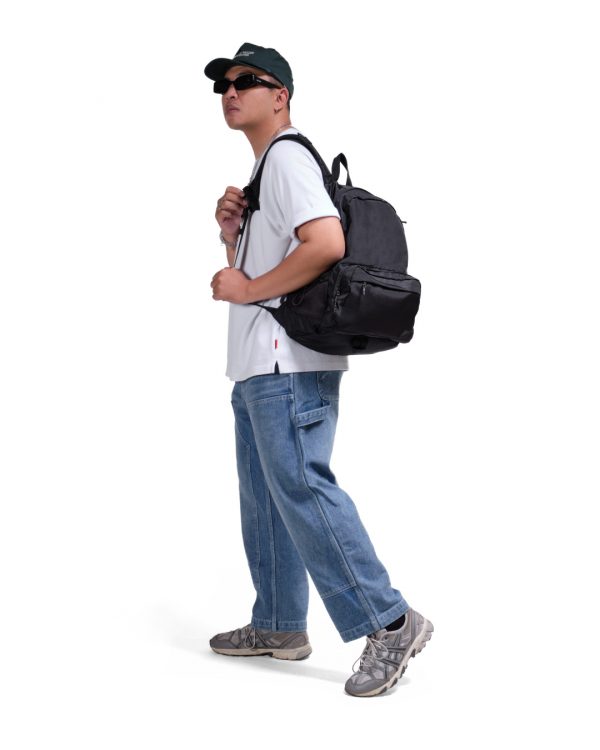 Full-body side view of a male model in urban streetwear wearing the black SYMA Foyer, demonstrating its large 21L scale and ergonomic fit. 16 inch commuter backpack.