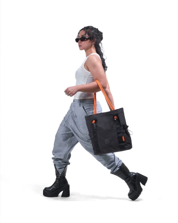 Full-body profile of a female model in modern streetwear carrying the black SYMA Noble minimalist everyday tote bag featuring contrasting orange paracord straps