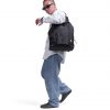 Full-body lifestyle shot of a male model carrying the black SYMA Chorus 17L 2-way backpack tote over one shoulder, highlighting its urban aesthetic, front utility pocket, and side mesh bottle pockets