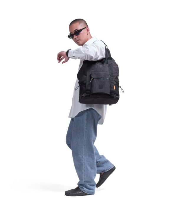 Full-body lifestyle shot of a male model carrying the black SYMA Chorus 17L 2-way backpack tote over one shoulder, highlighting its urban aesthetic, front utility pocket, and side mesh bottle pockets