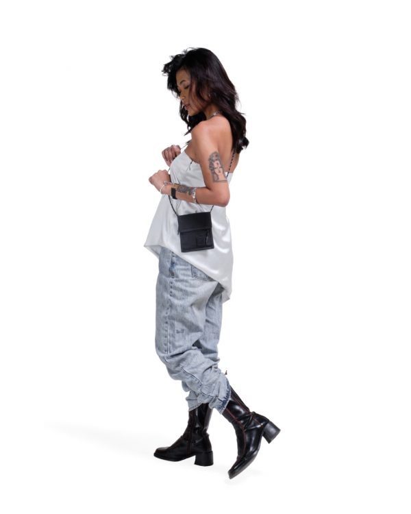 Female model in urban streetwear showcasing the black SYMA Arrival compact travel wallet worn as a hanging neck pouch bag over a white silk top and denim jeans