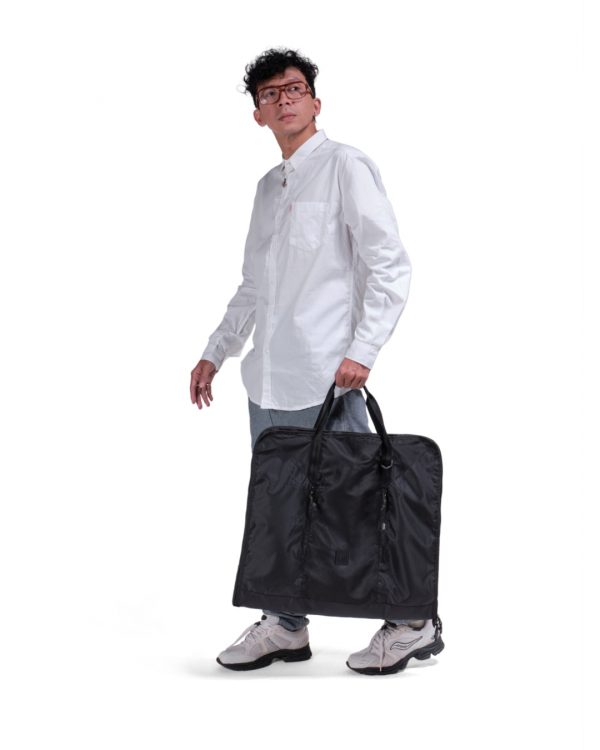 Male model in smart casual attire carrying the folded black SYMA Motion by its top handles, demonstrating its easy portability. Travel garment bag.