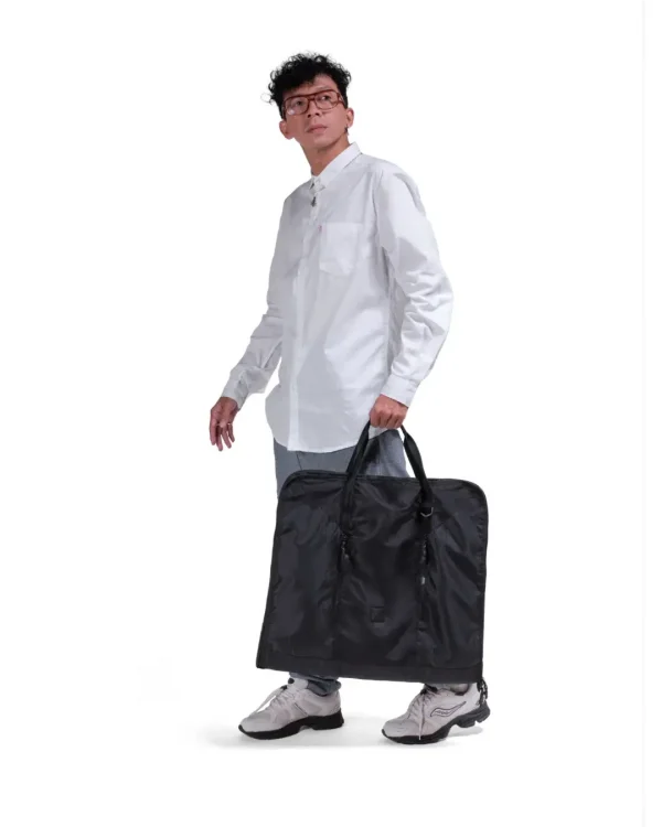 Male model in smart casual attire carrying the folded black SYMA Motion by its top handles, demonstrating its easy portability. Travel garment bag.