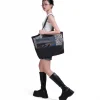 Side view of a female model in urban fashion carrying the black SYMA Peach on her shoulder, demonstrating its massive capacity and transparent mesh upper. Large urban tote bag.