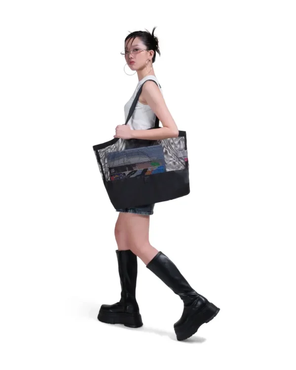 Side view of a female model in urban fashion carrying the black SYMA Peach on her shoulder, demonstrating its massive capacity and transparent mesh upper. Large urban tote bag.