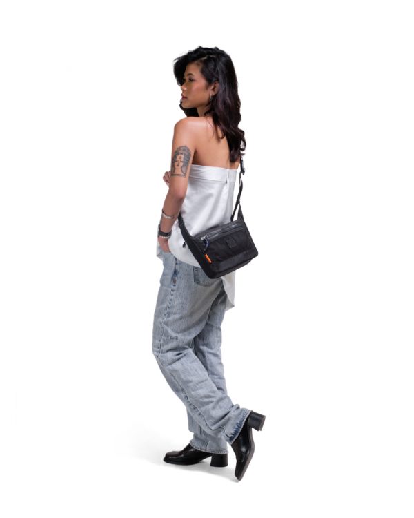 Full-body side view of a female model in casual streetwear wearing the black SYMA Strings Sling Bag, demonstrating its compact scale and front mesh pocket. Small urban crossbody bag.