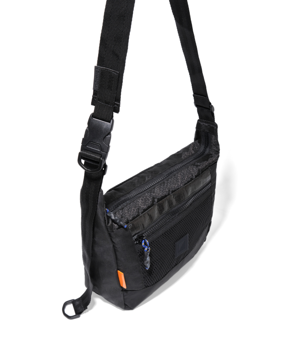 High-angle suspended view of the black SYMA Strings Sling Bag highlighting its tactical quick-release buckle, glossy PE Tarpaulin top panel, and zippered front mesh pocket. Small urban crossbody bag.