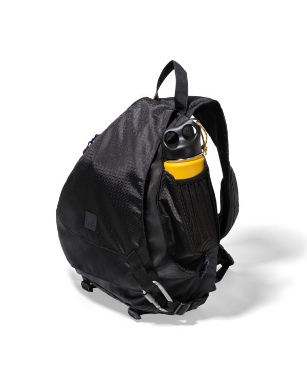 Angled side view of the black SYMA Symmetry Crossbody Bag resting on a white surface, highlighting its expandable side mesh pocket holding a yellow water bottle and hexagon ripstop exterior. Large techwear crossbody bag.