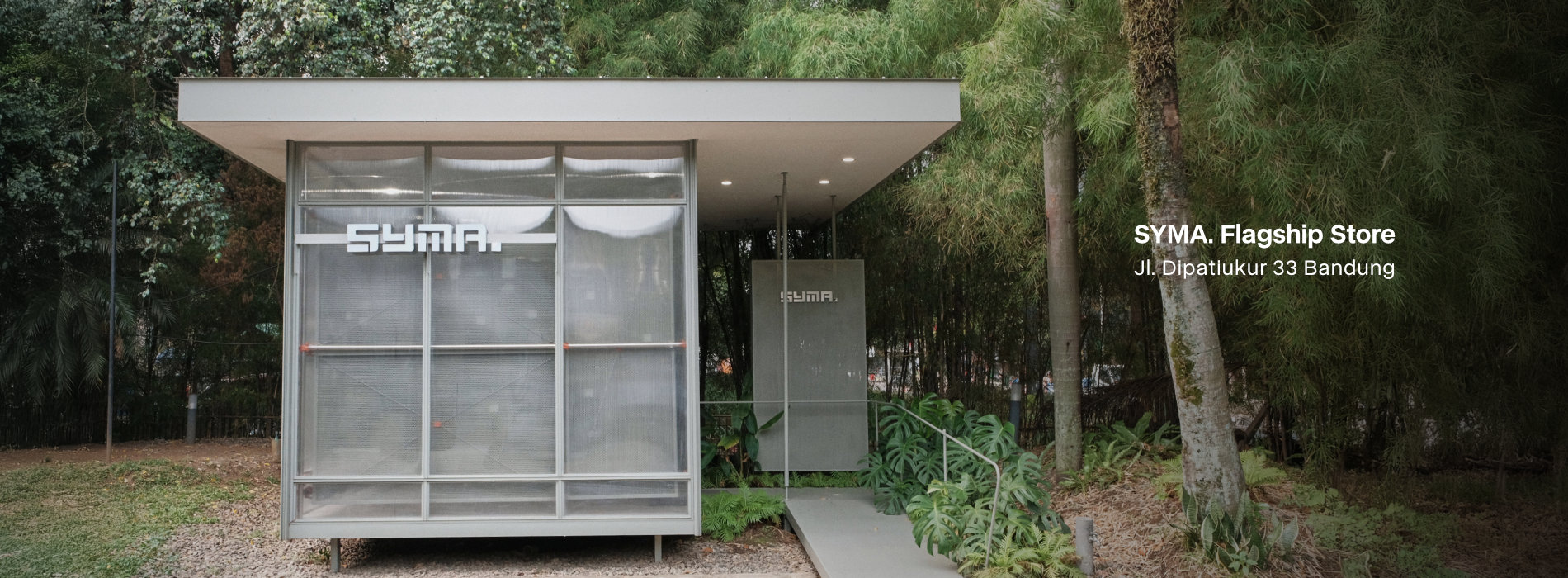 Desktop website banner showcasing the physical SYMA Flagship Store, featuring a modern translucent minimalist building surrounded by green bamboo trees, with text reading: SYMA. Flagship Store, Jl. Dipatiukur 33 Bandung