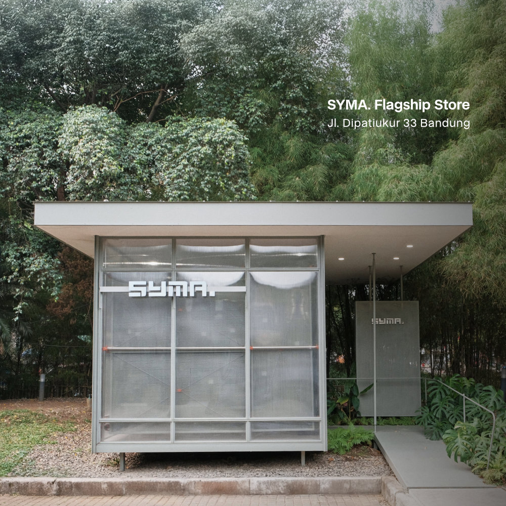 Mobile website banner showcasing the physical SYMA Flagship Store, featuring a modern translucent minimalist building surrounded by lush green trees, with text reading: SYMA. Flagship Store, Jl. Dipatiukur 33 Bandung