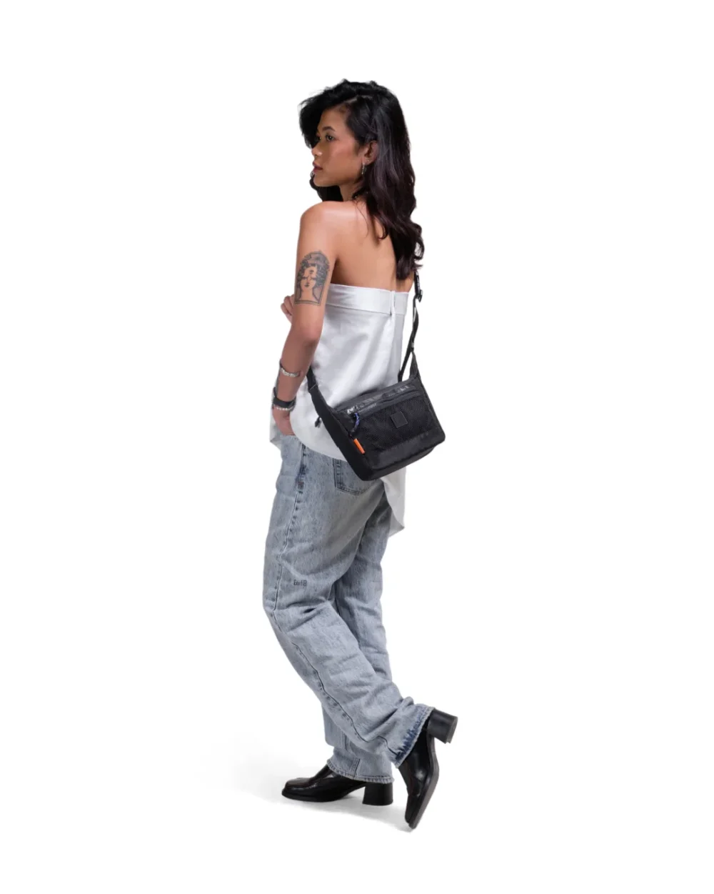 Full-body side view of a female model in casual streetwear wearing the black SYMA Strings Sling Bag, demonstrating its compact scale and front mesh pocket. Small urban crossbody bag.