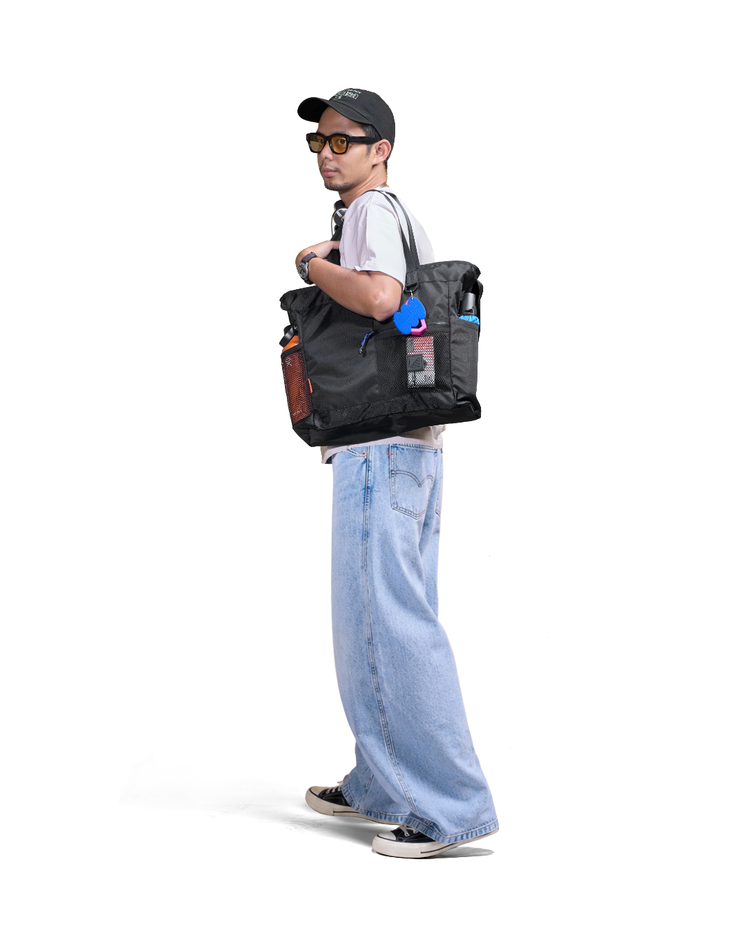 Side profile view of a male model in casual urban streetwear carrying the black SYMA Bridge Tote Bag on his shoulder, showcasing the premium urban utility tote's side mesh pockets, blue paracord zipper pull, and a vibrant blue and pink SYMA bag charm attached to the D-ring