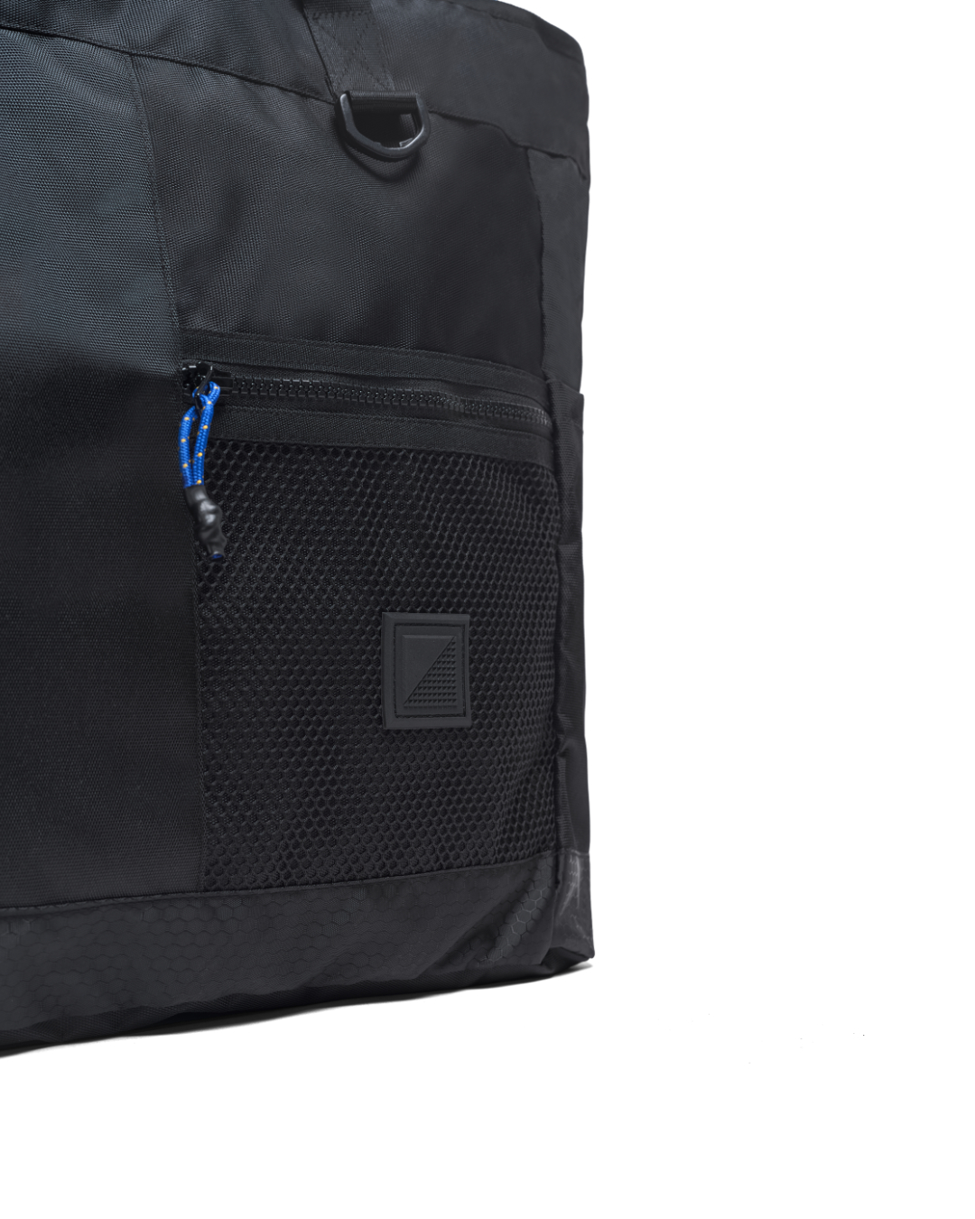 Extreme close-up detail of the black SYMA Bridge Tote Bag, highlighting the premium water-resistant nylon, a front mesh pocket with a custom blue paracord YKK zipper pull, a matte black D-ring, and the tonal black SYMA logo patch